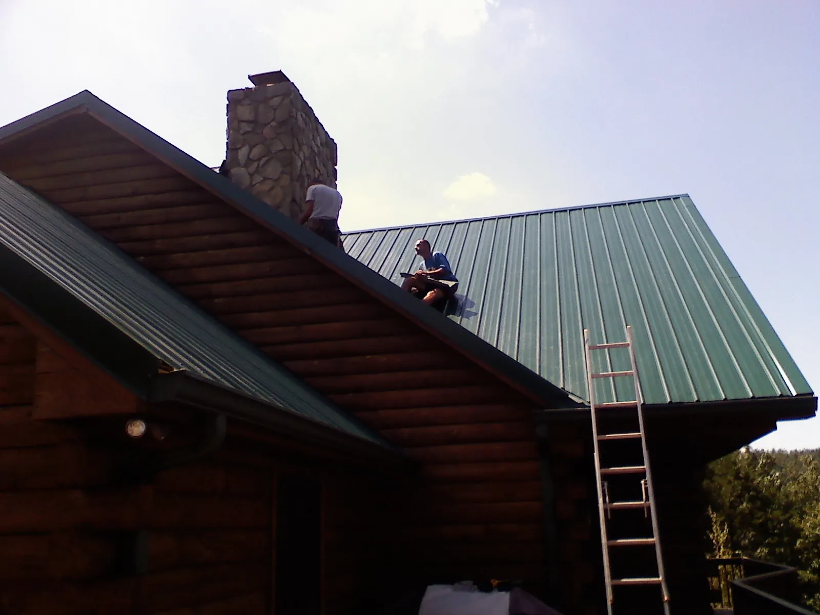 Roofers installing green metal panels beside a stone chimney on a log home in Verona